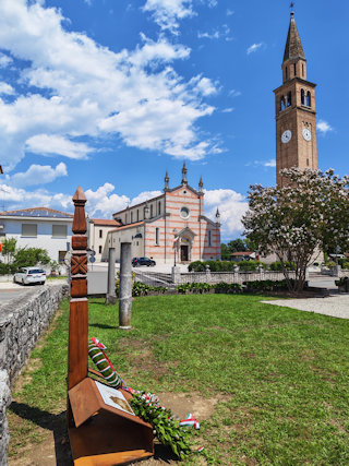 A Király Ivánra és az olasz‒magyar barátságra emlékeztető kopjafa a háttérben a gyertyatartót őrző Falzè di Piave-i római katolikus templommal.